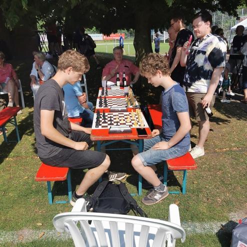 Zwei Jungen spielen Schach an einem Tisch im Freien, umgeben von Zuschauern und Bäumen.