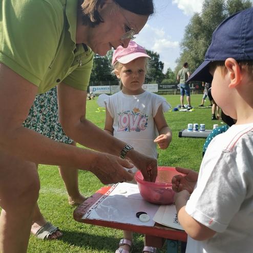Frau hilft zwei Kindern beim Spielen mit einer bunten Schüssel auf einem Tisch im Freien.