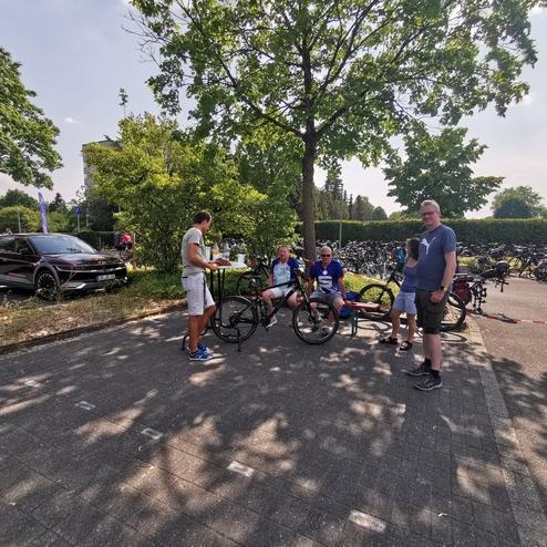 Gruppengespräch von vier Personen mit Fahrrädern vor einem Fahrradparkplatz und einem schwarzen Auto im Hintergrund.