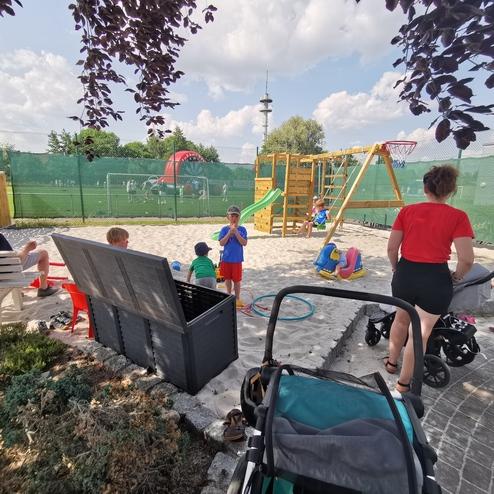 Spielplatz mit Sandfläche, Kindern, Spielgeräten und einer schauenden Erwachsenen mit Kinderwagen im Hintergrund.