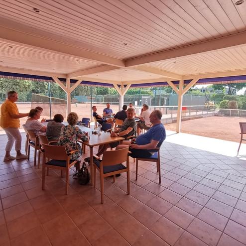 Gruppe von Menschen sitzt an einem Tisch unter einer überdachten Terrasse neben Tennisplätzen. Ein Gespräch findet statt.