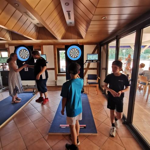Vier Kinder und ein Erwachsener interagieren in einem Dartsalon mit elektronischen Dartscheiben und Bildschirmen.