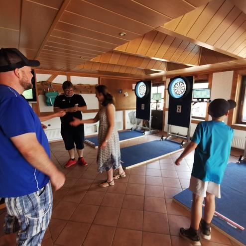 Gruppe von vier Personen in einem Dartraum, einige spielen, während andere zuschauen. Dartboards im Hintergrund.