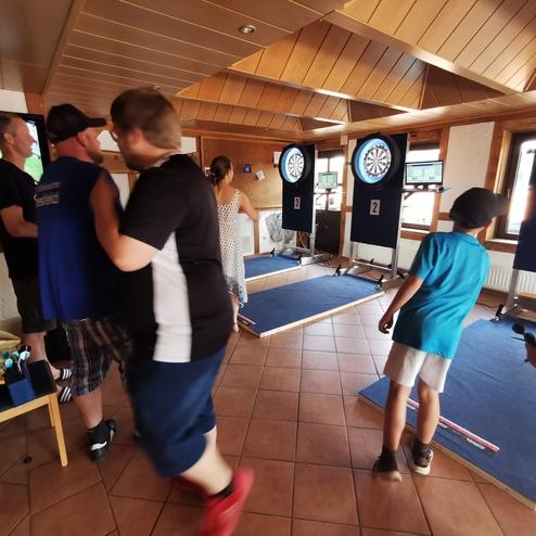 Gruppenszene in einem Dartraum mit mehreren Personen und drei Dartboards im Hintergrund. Bildschirm zeigt Sportübertragung.