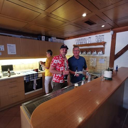 Zwei Männer stehen an der Bar in einer Küche, einer hält ein Bier und der andere ein Glas. Im Hintergrund arbeitet eine Person.