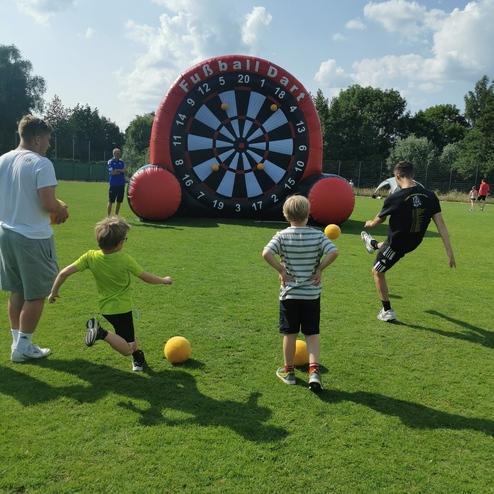 Kinder spielen Fußball-Dart mit einem großen, aufblasbaren Ziel auf einem grünen Sportplatz unter blauem Himmel.