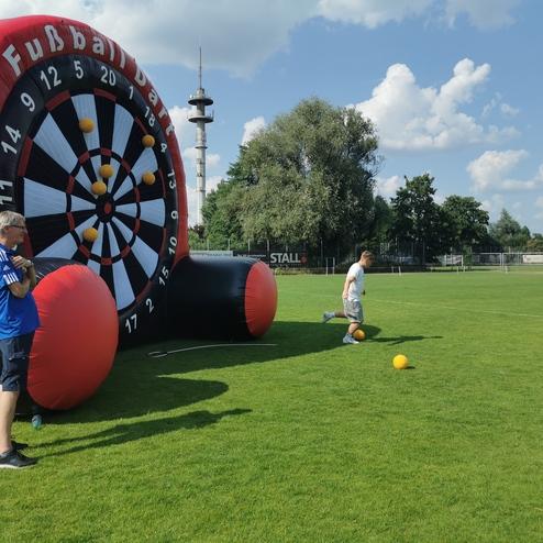 Mann tritt einen Ball Richtung aufblasbares Fußball-Dartspiel auf einem Rasenplatz, während ein Zuschauer zuschaut.