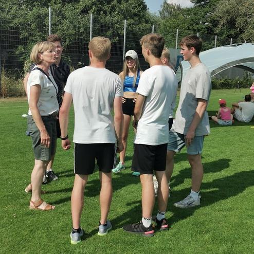 Gruppe von sieben Jugendlichen in sportlicher Kleidung steht auf einer Wiese und unterhält sich mit einer erwachsenen Frau.
