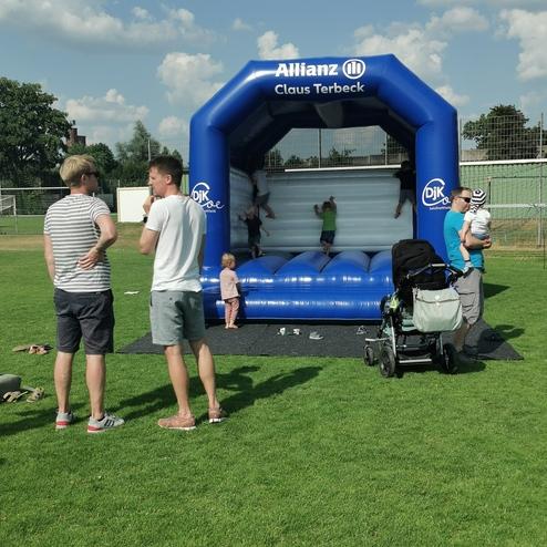 Gruppen von Menschen stehen auf einem Rasen neben einer blauen Hüpfburg mit Kindern, die darin springen und spielen.
