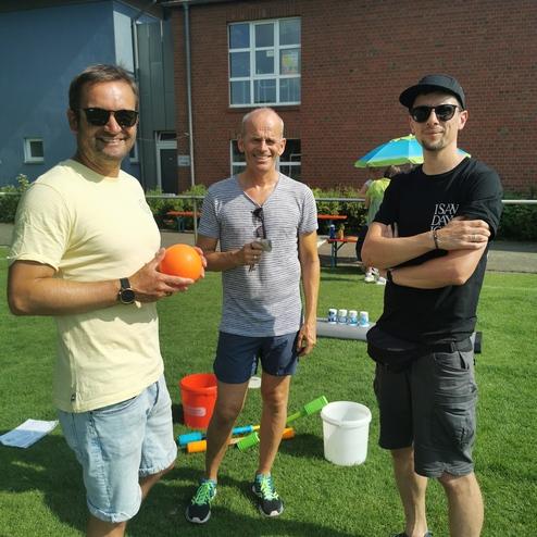 Drei Männer stehen auf einer Wiese, einer hält einen orangefarbenen Ball, im Hintergrund sind Spielutensilien sichtbar.