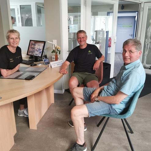 Drei Personen sitzen in einem Empfangsbereich, während eine Mitarbeiterin hinter einem Schreibtisch arbeitet.