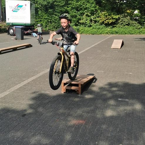 Junge springt mit Fahrrad über eine Holzrampe auf einem Parkplatz, im Hintergrund weitere Kinder auf Fahrrädern.