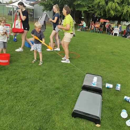 Kinder spielen im Freien mit Wasser und Schläuchen, während Erwachsene mit Handtüchern und Spielmaterialien beschäftigt sind.
