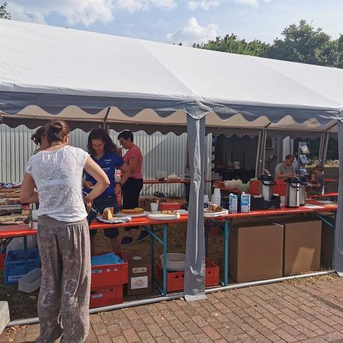 Personen stehen an einem Tisch unter einem Zelt und bereiten Essen und Getränke für eine Veranstaltung vor.