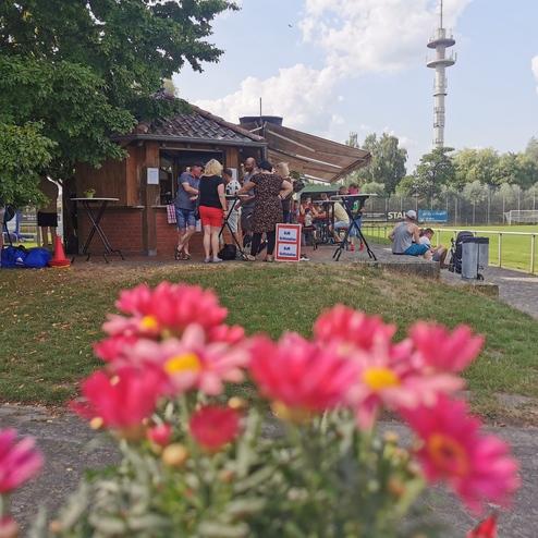 Gruppierung von Personen vor einem Vereinsheim, im Vordergrund rosa Blumen, im Hintergrund ein Fußballfeld und Zuschauer.