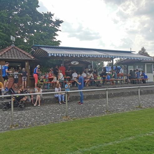 Zahlreiche Menschen sitzen und stehen vor einem Kiosk mit überdachten Tischen auf einer Sportanlage.