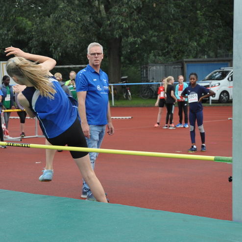 Mädchen springt im Hochsprung über eine Latte, während Trainer und andere Athleten auf dem Sportplatz zuschauen.