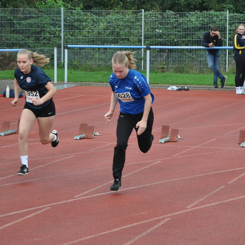 Zwei junge Läuferinnen sprinten auf einer roten Bahn, während Zuschauer im Hintergrund stehen.