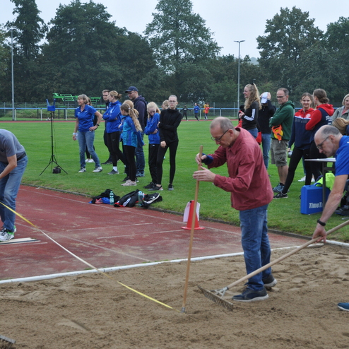 Drei Männer messen und bearbeiten eine Sandgrube auf einem Sportplatz, während Zuschauer im Hintergrund beobachten.
