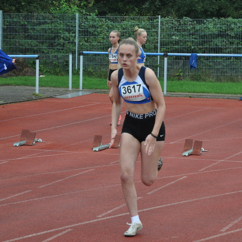 Eine Athletin sprintet auf einer roten Laufbahn, während andere Teilnehmerinnen im Hintergrund bereitstehen.
