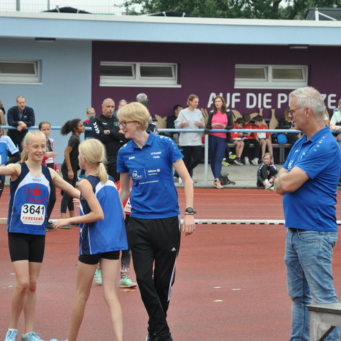 Gruppierung von jungen Leichtathleten in blauen Trikots, mit Betreuern auf einer roten Laufbahn vor Zuschauern.