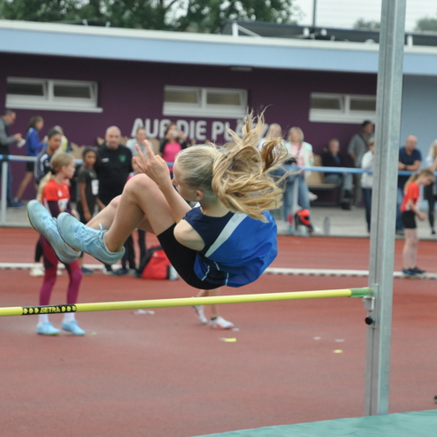 Mädchen springt über eine Hochsprungstange auf einem Sportplatz, Zuschauer im Hintergrund beobachten den Wettkampf.