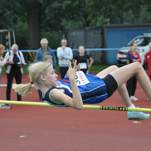 Mädchen springt über eine Hochsprunglatte auf einem Sportplatz, während Zuschauer im Hintergrund zuschauen.