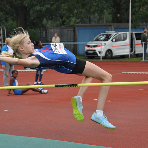 Junge Athletin springt &uuml;ber eine Hochsprungstange auf einer Laufbahn, umgeben von anderen Sportlern und Zuschauern.