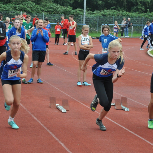 Zwei junge Athletinnen starten sprintend von Blocks auf einer roten Laufbahn, während Zuschauer im Hintergrund jubeln.