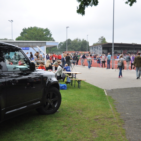 Schwarzer SUV im Vordergrund, im Hintergrund zahlreiche Menschen bei einer Sportveranstaltung auf einer Laufbahn.
