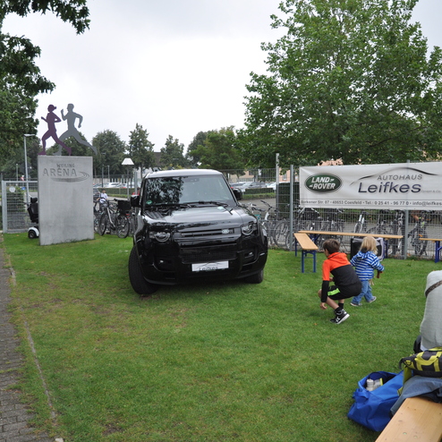 Schwarzer SUV parkt auf Rasen neben dem Eingang zur Weiling Arena, umgeben von spielenden Kindern und Fahrrädern.