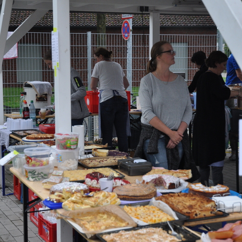 Stand mit verschiedenen Kuchen und Snacks, umgeben von Personen bei einem Verkaufsstand unter einem Pavillon.