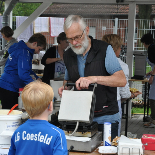 Älterer Mann mit Brille macht Waffeln an einer Verkaufsbude, ein Junge schaut interessiert zu. Hintergründe mit weiteren Helfern.