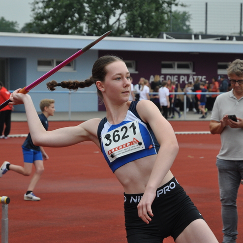 Sportlerin wirft einen Speer auf einem Leichtathletikfeld, im Hintergrund Zuschauer und andere Athleten.