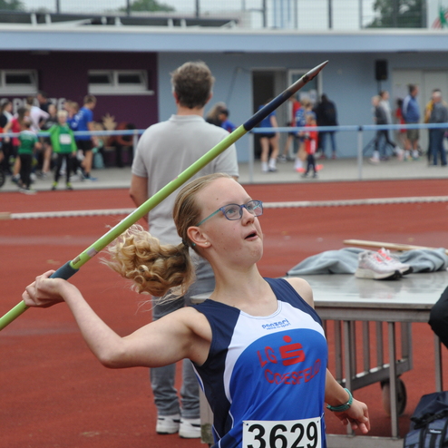 M&auml;dchen wirft einen Speer auf einer Sportveranstaltung, im Hintergrund Zuschauer und andere Athleten.