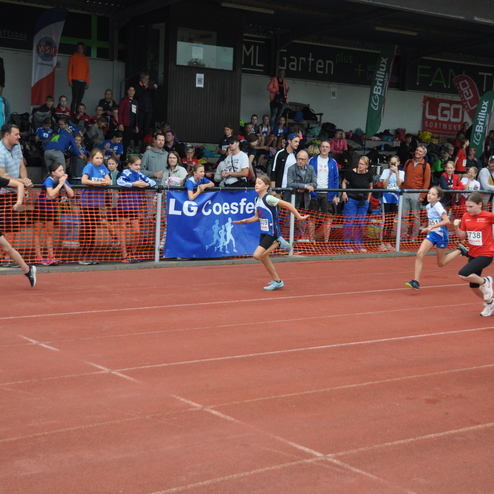 Vier junge Leichtathleten sprinten auf einer roten Laufbahn w&auml;hrend eines Wettkampfs, Zuschauer stehen auf der Trib&uuml;ne.