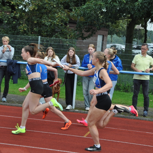 Zwei Athletinnen &uuml;bergeben den Staffelstab auf einer roten Laufbahn, im Hintergrund zuschauende Personen.
