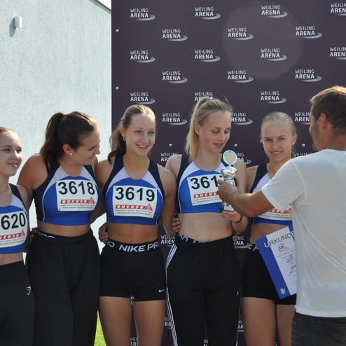Sechs junge Athletinnen stehen beim Wettkampf zusammen und erhalten einen Pokal von einem Mann in einem wei&szlig;en T-Shirt.