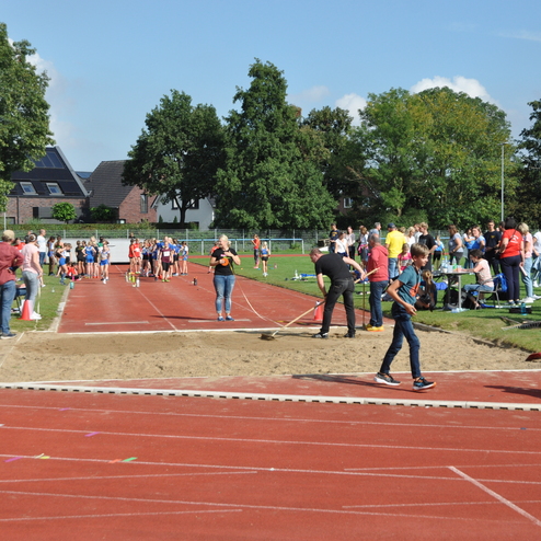 Sportveranstaltung im Freien mit Kindern, Zuschauern und einem Weitsprungbereich auf einer roten Tartanbahn.