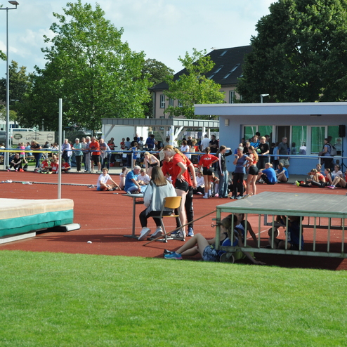 Sportveranstaltung auf einem Au&szlig;ensportplatz mit Teilnehmern und Zuschauern, im Hintergrund eine Trib&uuml;ne und B&auml;ume.
