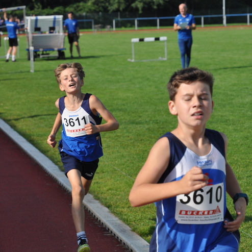Zwei Kinder in blauen Trikots laufen auf einer Wettkampfbahn; im Hintergrund weitere Sportler und Zuschauer.