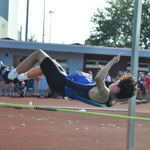 Ein Hochspringer in blauer Sportkleidung &uuml;berquert eine Stange bei einem Wettkampf auf einer roten Bahn, Zuschauer im Hintergrund.