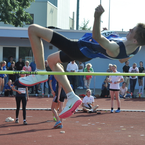 Ein Hochspringer in blauem Trikot &uuml;berspringt eine Latte bei einem Wettkampf, Zuschauer sitzen im Hintergrund.