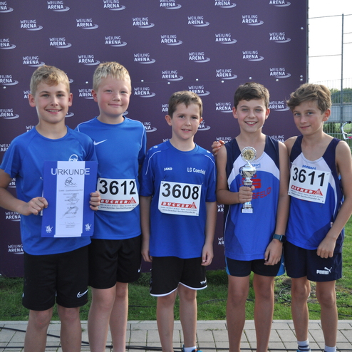 F&uuml;nf junge Leichtathleten posieren mit Urkunde und Pokal vor einer Sponsorenwand nach einem Wettkampf.