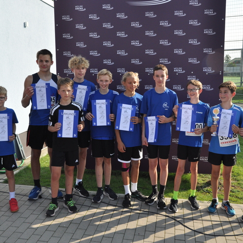 Gruppe von Jungen in sportlichen Outfits, die Urkunden und einen Pokal vor einer Eventwand halten.