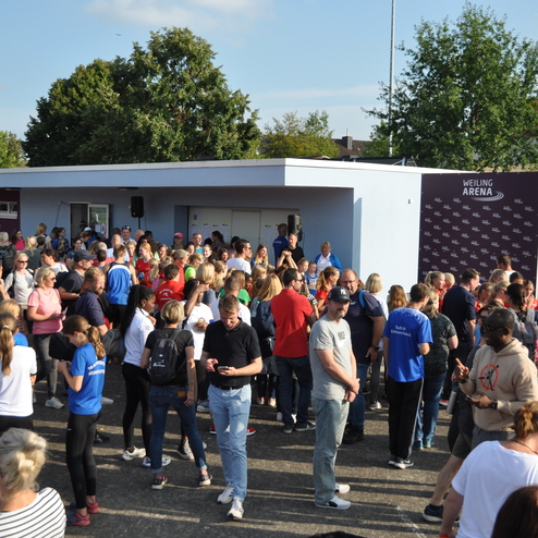 Menschenmenge versammelt sich vor dem Eingang der Weiling Arena, umgeben von B&auml;umen und hellblauem Himmel.