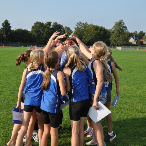 Gruppierung von M&auml;dchen in blauen Sportanz&uuml;gen, die sich um einen Pokal auf einem Rasenplatz versammeln.