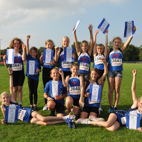 Gruppe junger Athletinnen mit Urkunden l&auml;chelt auf gr&uuml;nem Sportplatz, einige halten die Urkunden hoch.