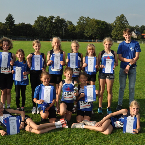 Gruppe von Jugendlichen mit Urkunden auf einer Sportwiese, betreut von einer Trainerin im blauen Shirt.
