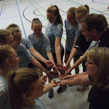 Gruppe von acht Mädchen und einem Trainer, die sich in einer Sporthalle zum Teamgeist motivieren und ihre Hände zusammenbringen.